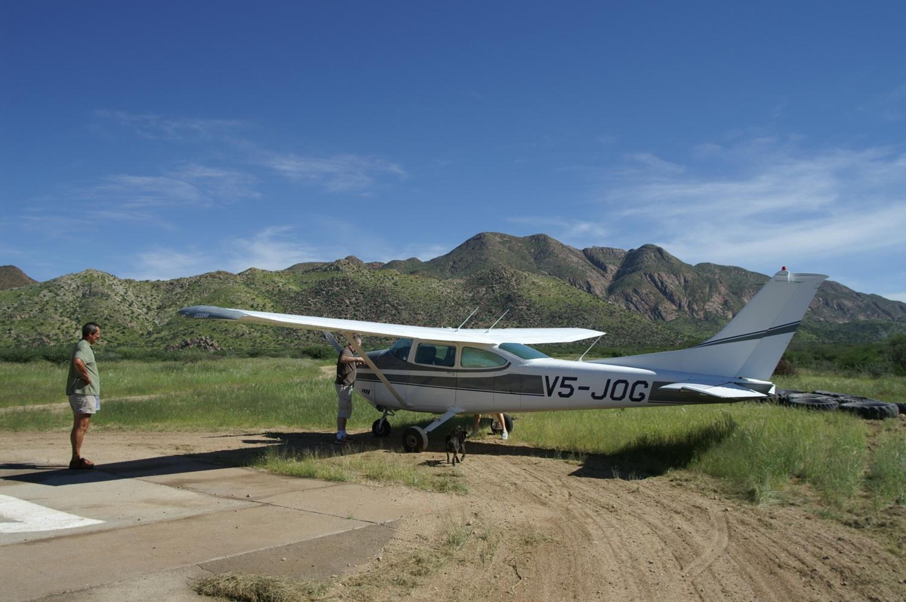 Una aeronave pequeña con el código de identificación V5-JOG estacionada sobre una superficie de tierra, con montañas al fondo.