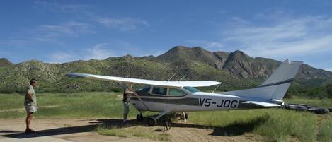 Una aeronave pequeña con el código de identificación V5-JOG estacionada sobre una superficie de tierra, con montañas al fondo.
