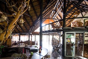 Un interior rústico con techo alto, vigas de madera y techo de paja. Hay mobiliario de mimbre y un ventanal de vidrio que da a otra habitación.