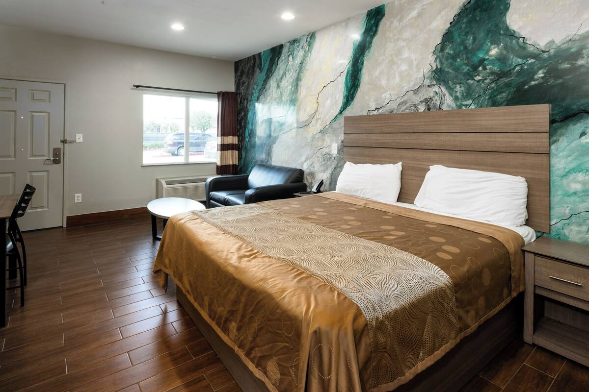 Quarto de hotel com uma cama grande, uma cadeira, uma mesinha e uma janela com cortinas.