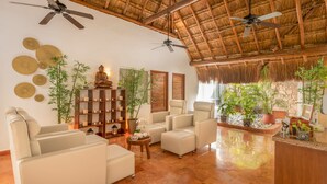 A spacious living area with a high ceiling, ceiling fans, a bamboo ceiling, and a variety of seating including sofas and armchairs. There are several potted plants and a small table with a teapot. - Desire Pearl Riviera Maya Resort – All Inclusive, Exclusively for Couples (Puerto Morelos)
