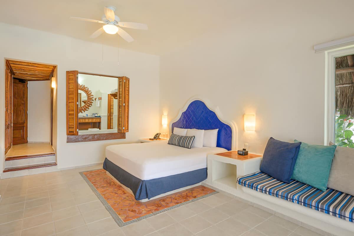 Habitación de hotel con una cama grande, una mesita de noche con lámpara, un rincón de descanso con cojines, un ventilador de techo y un espejo grande.