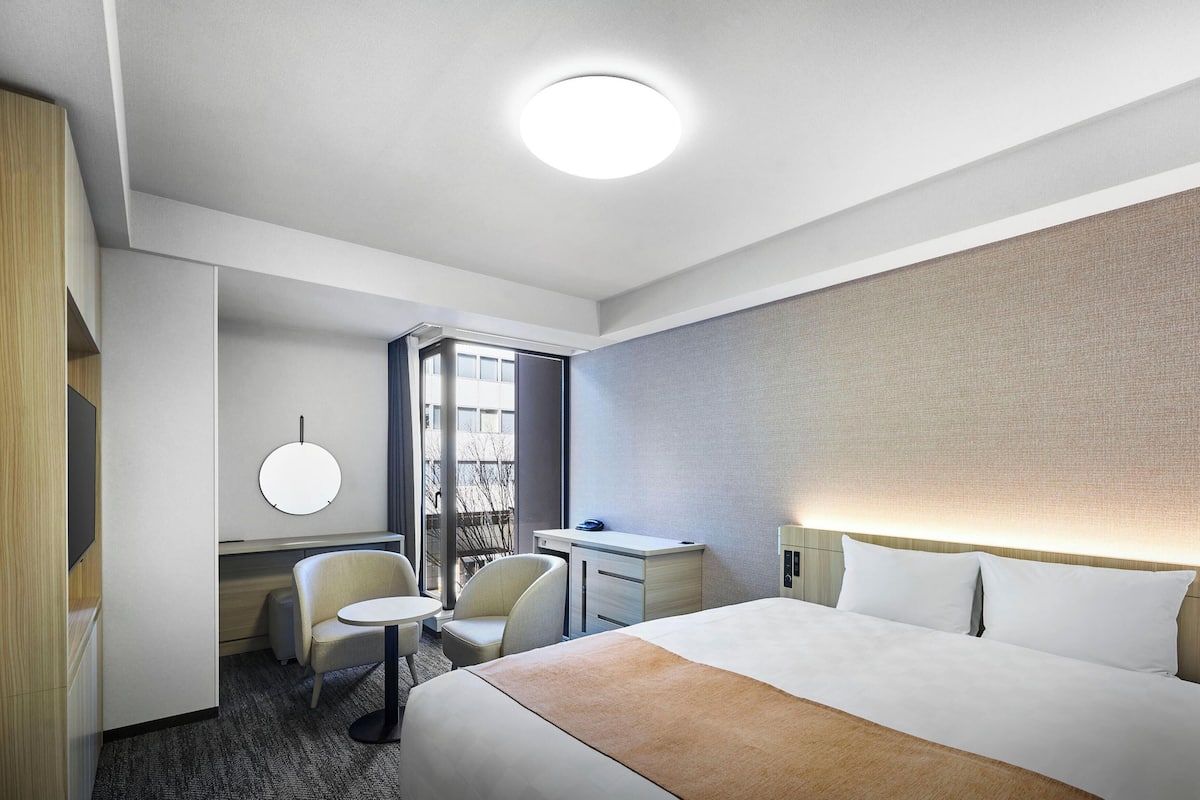 Une chambre d’hôtel avec un lit, un bureau, une chaise, un miroir rond et une fenêtre.
