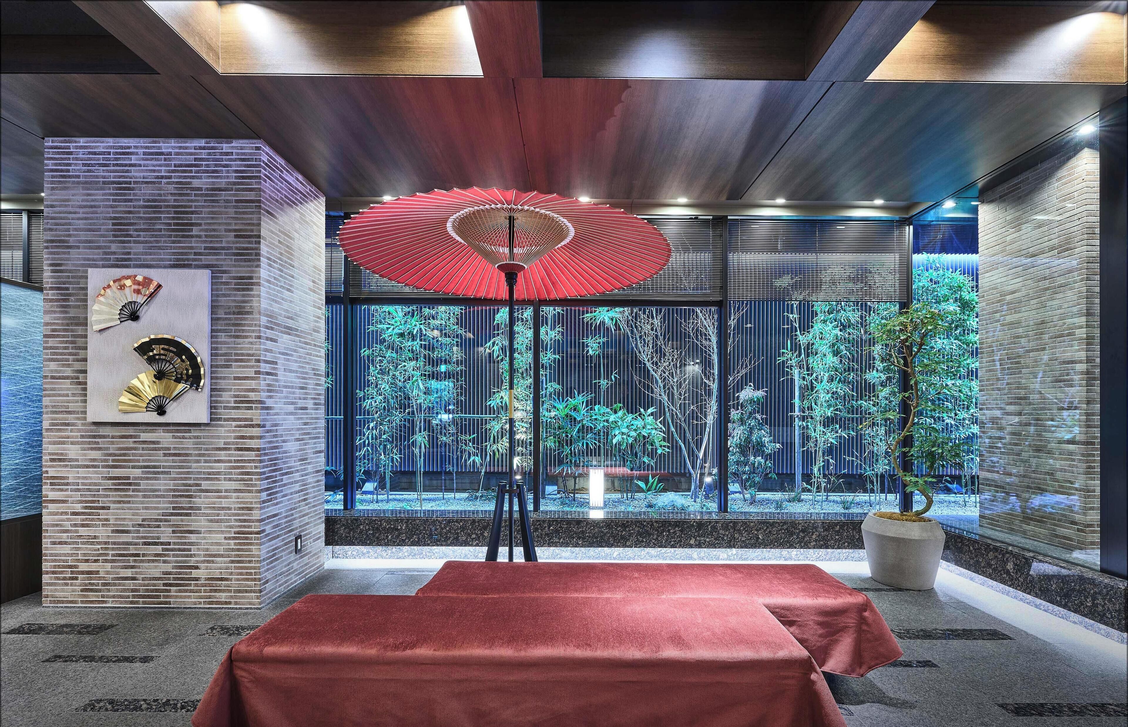 A room with a red umbrella, a fan, and a view of greenery through large windows.