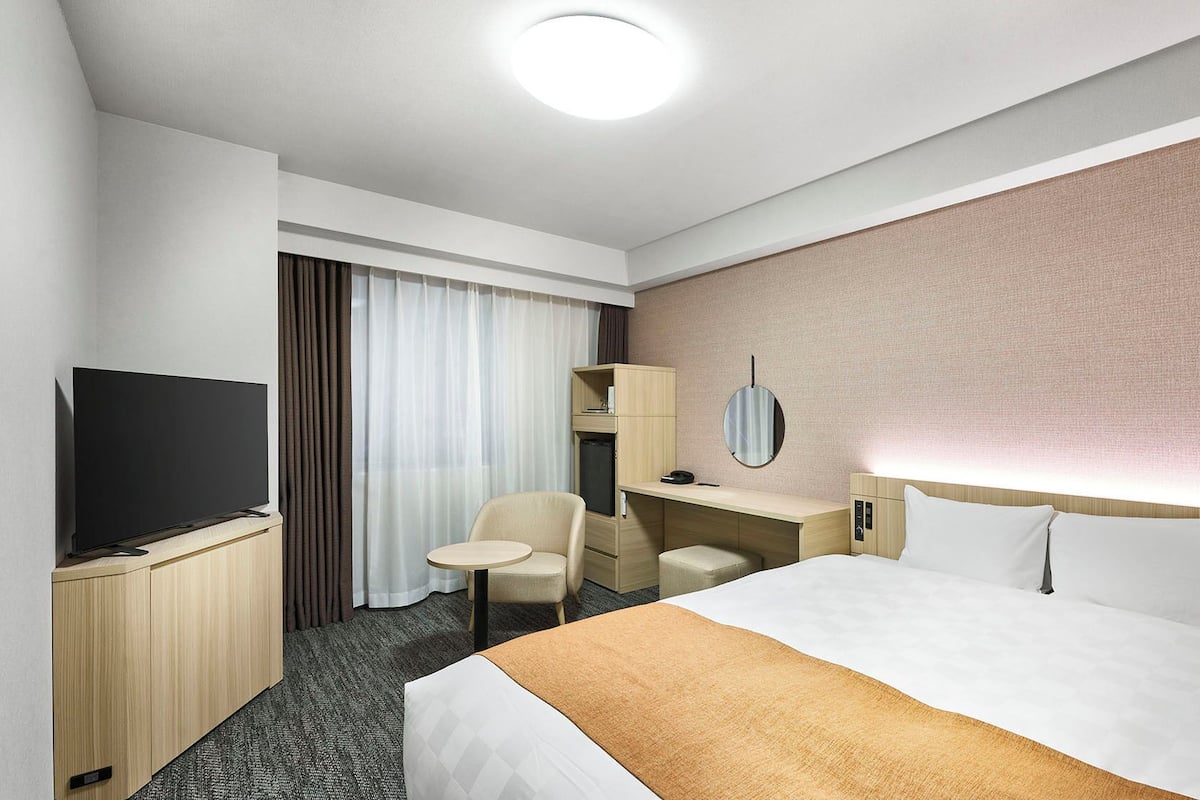 Une chambre d’hôtel équipée d’un lit, d’un bureau, d’une chaise, d’une télévision et d’une grande fenêtre avec des rideaux.