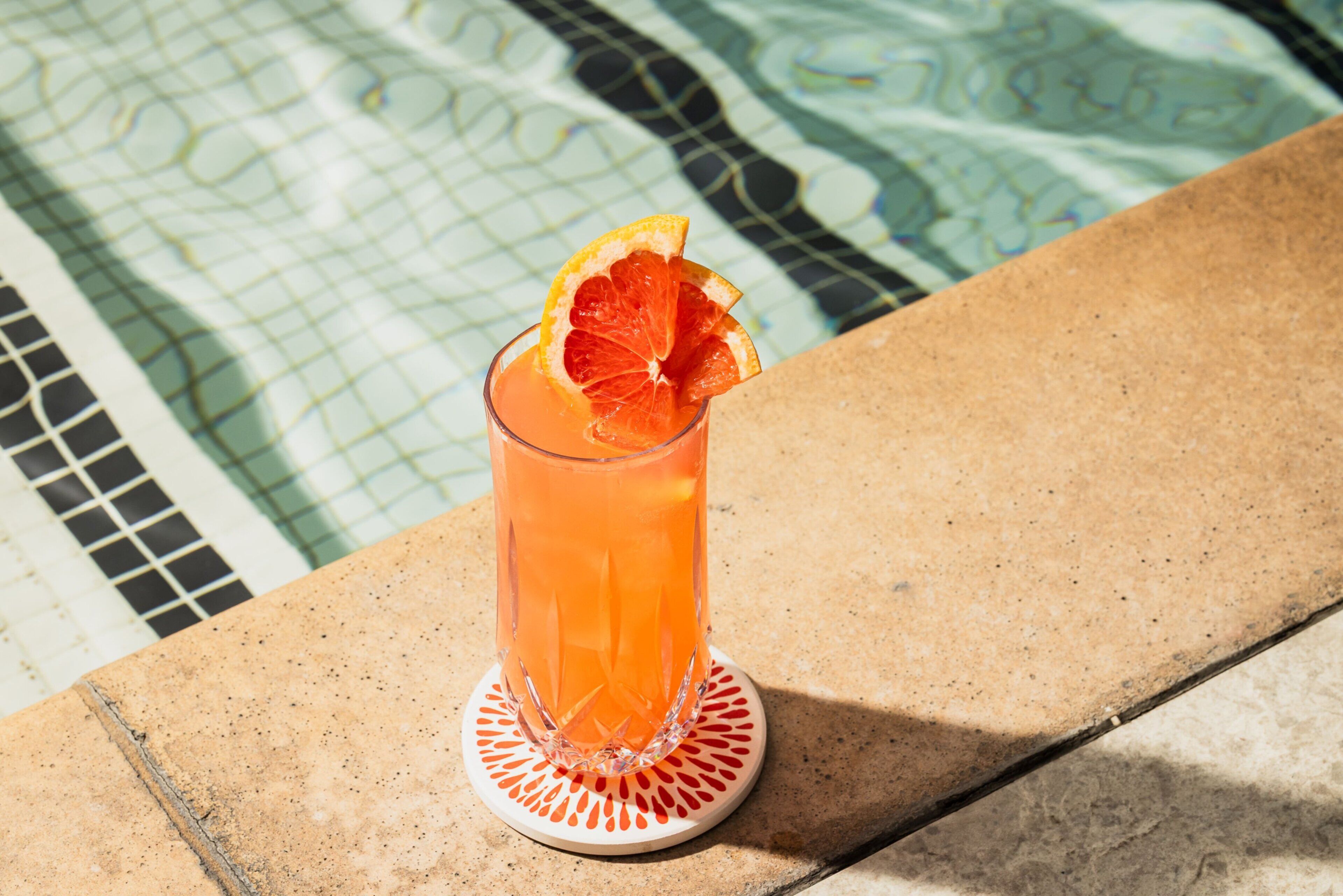 A glass of orange beverage with a slice of orange on a coaster by a pool.