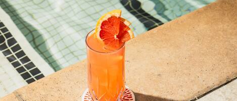 A glass of orange beverage with a slice of orange on a coaster by a pool.