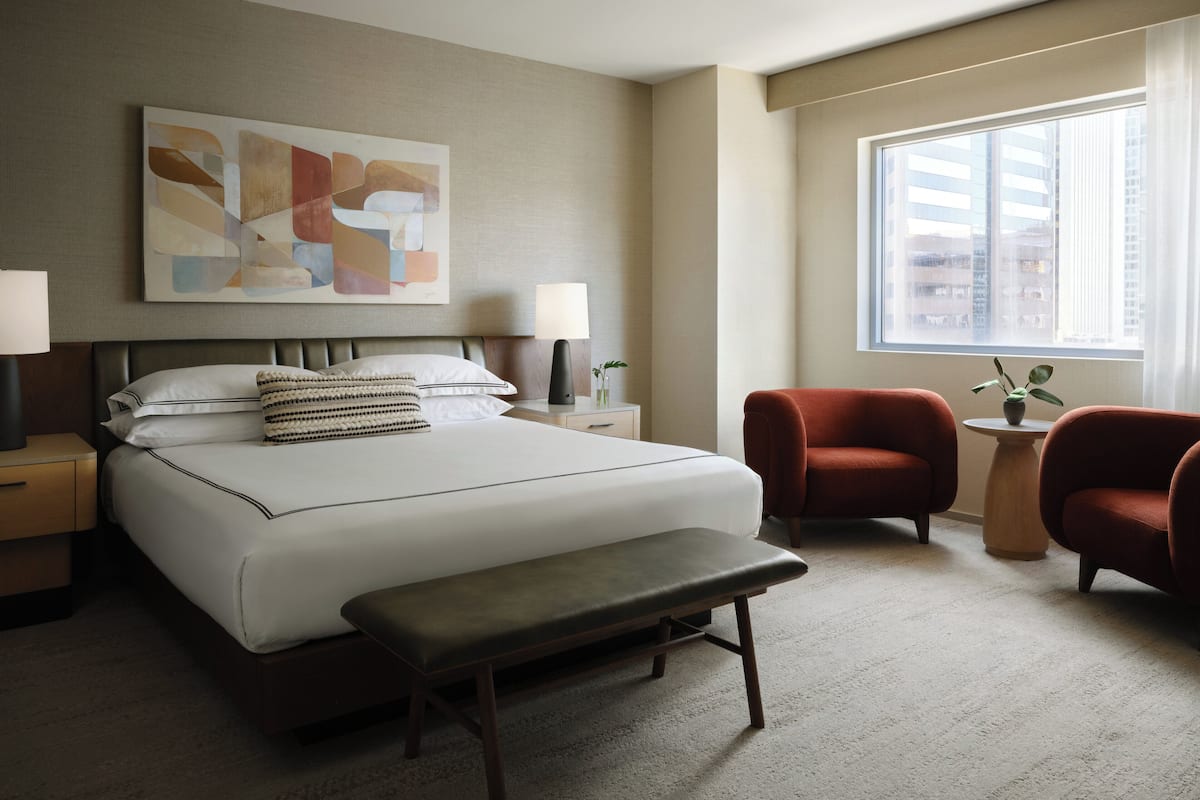 A hotel room with a large bed, a bench, two chairs, a nightstand with a lamp, a small table with a plant, and a window with a view of buildings.