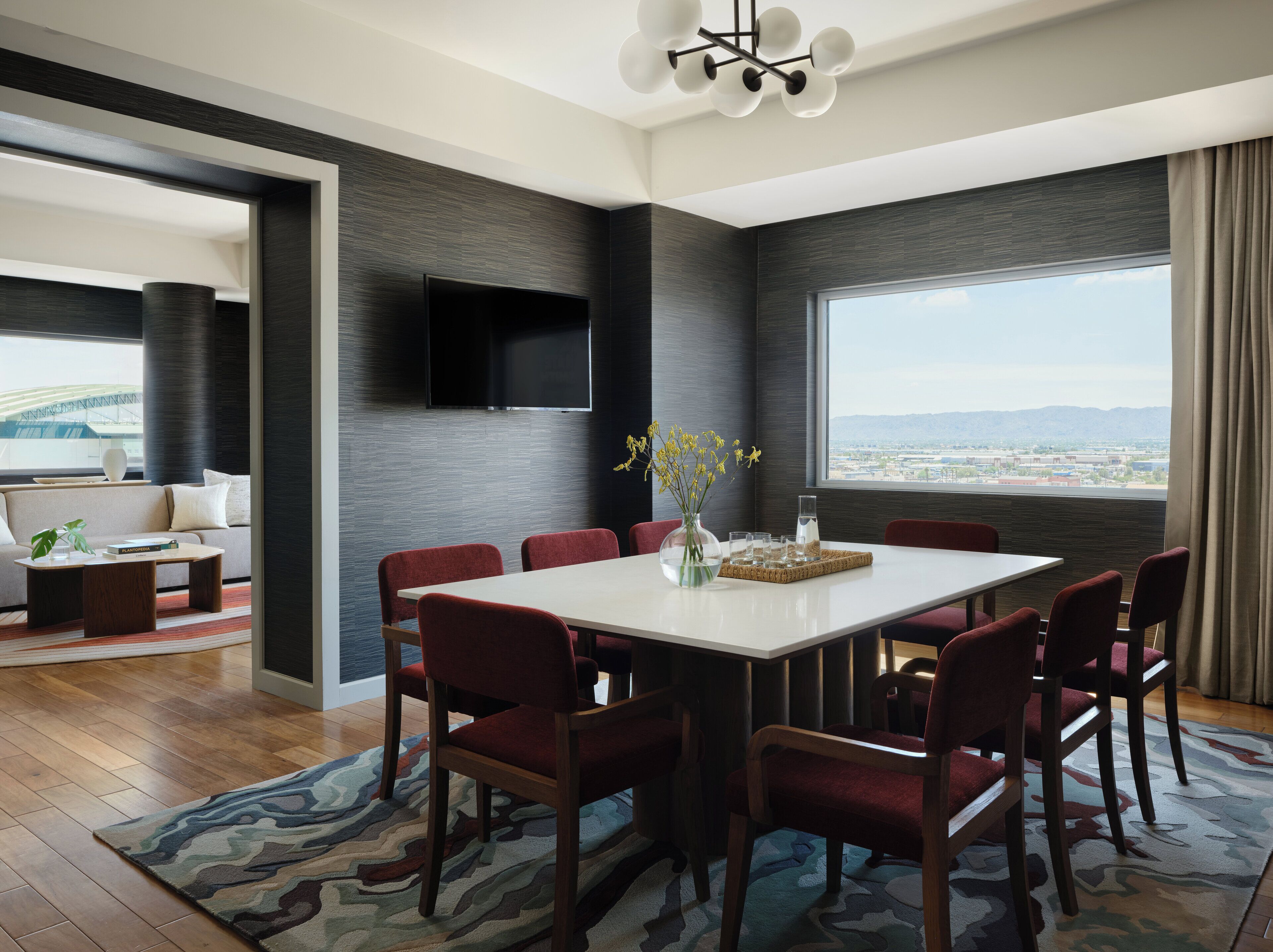 Un comedor moderno con una mesa blanca, sillas rojas y vistas a un paisaje urbano a través de amplios ventanales.