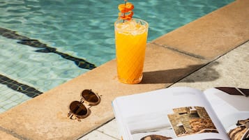 A glass of orange juice with a garnish, sunglasses, and an open magazine by a pool.
