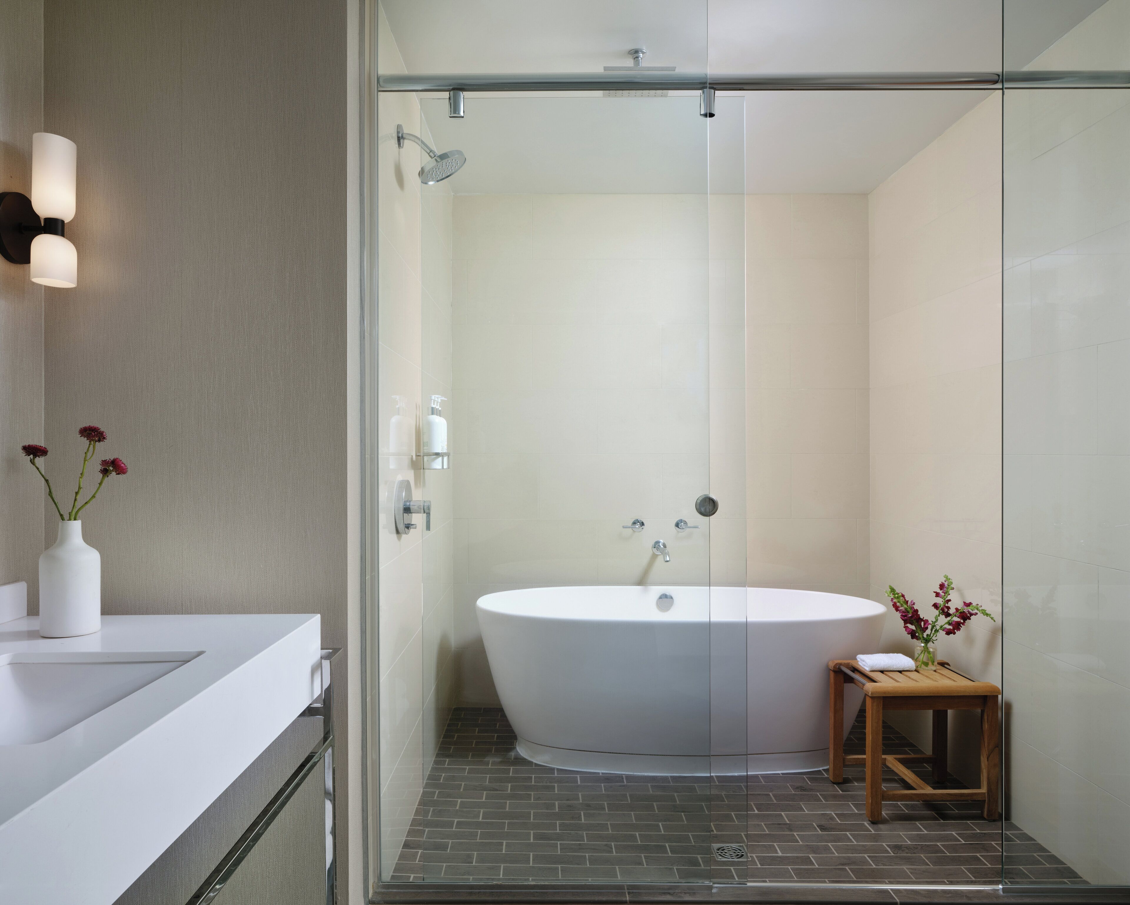 A modern bathroom with a glass-enclosed shower, a white bathtub, a wooden stool, and a vase with flowers on the counter.
