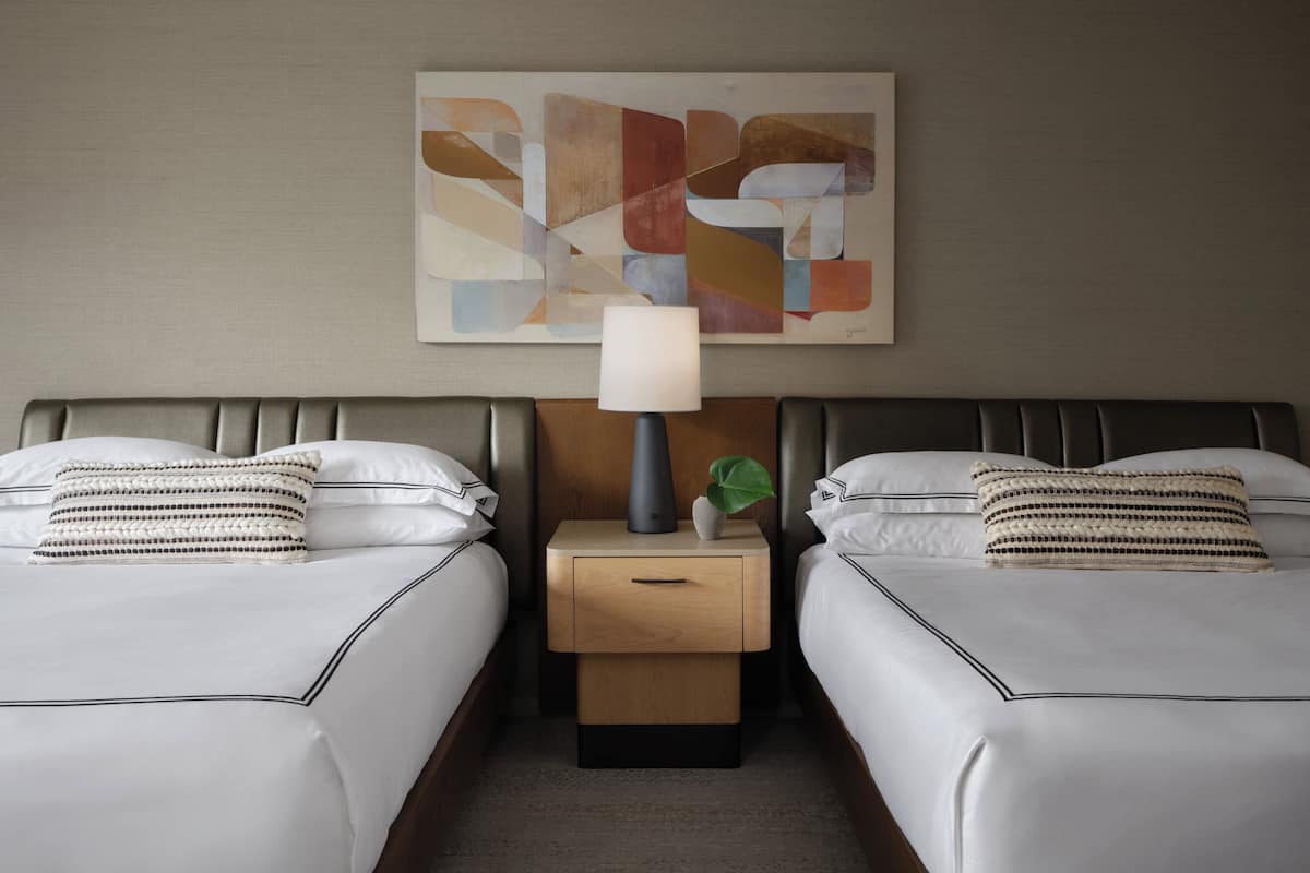 A hotel room with two beds, a nightstand with a lamp, a small plant, and a piece of abstract art on the wall.