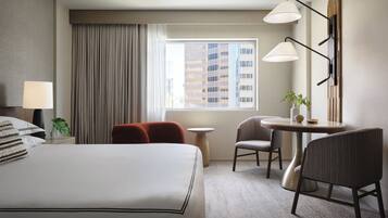 Habitación de hotel con cama, escritorio, dos sillas, una mesita, lámpara, planta y vista a un paisaje urbano por la ventana.