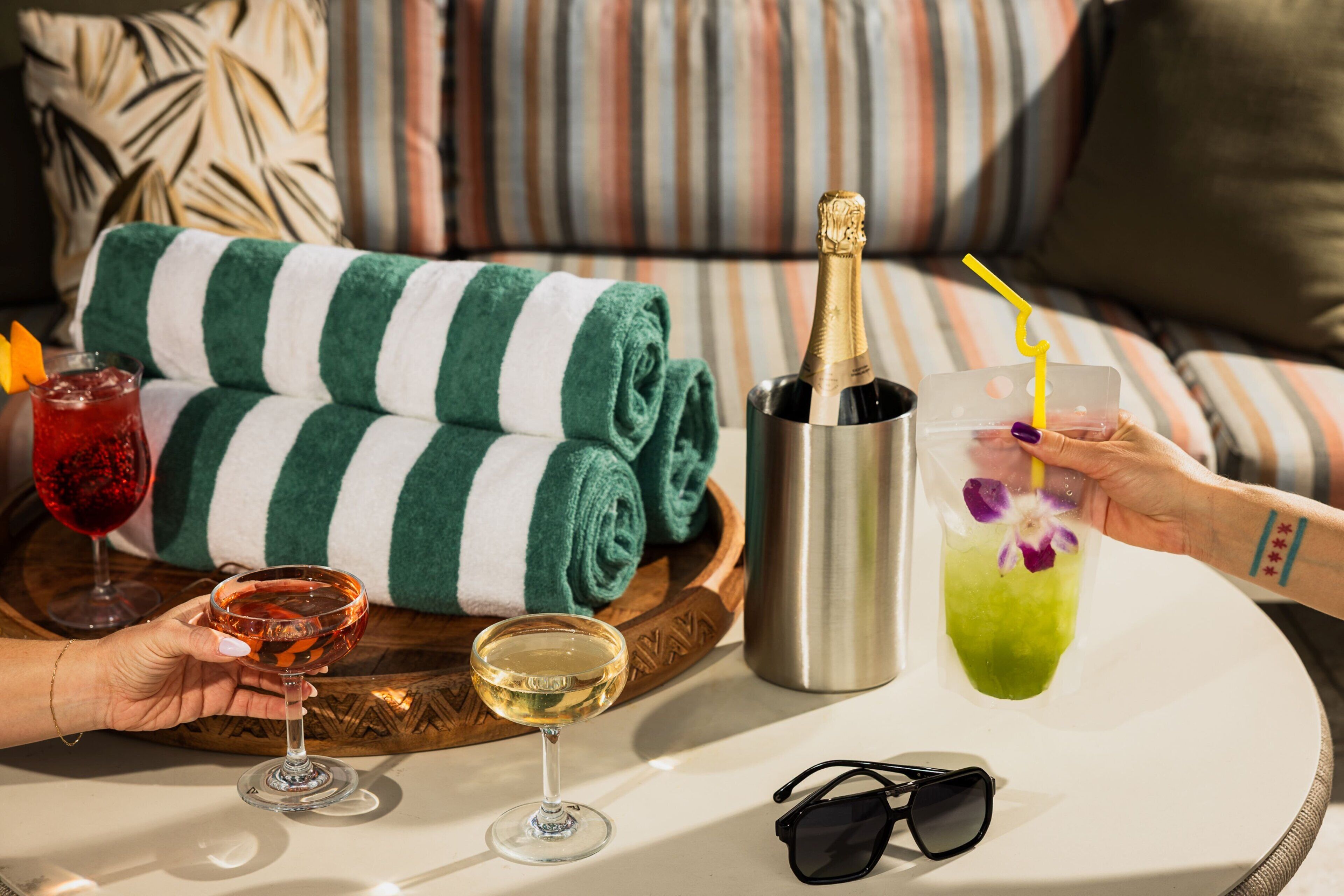 Una mesa con bebidas, una botella de champán, un par de gafas de sol y un cojín a rayas.