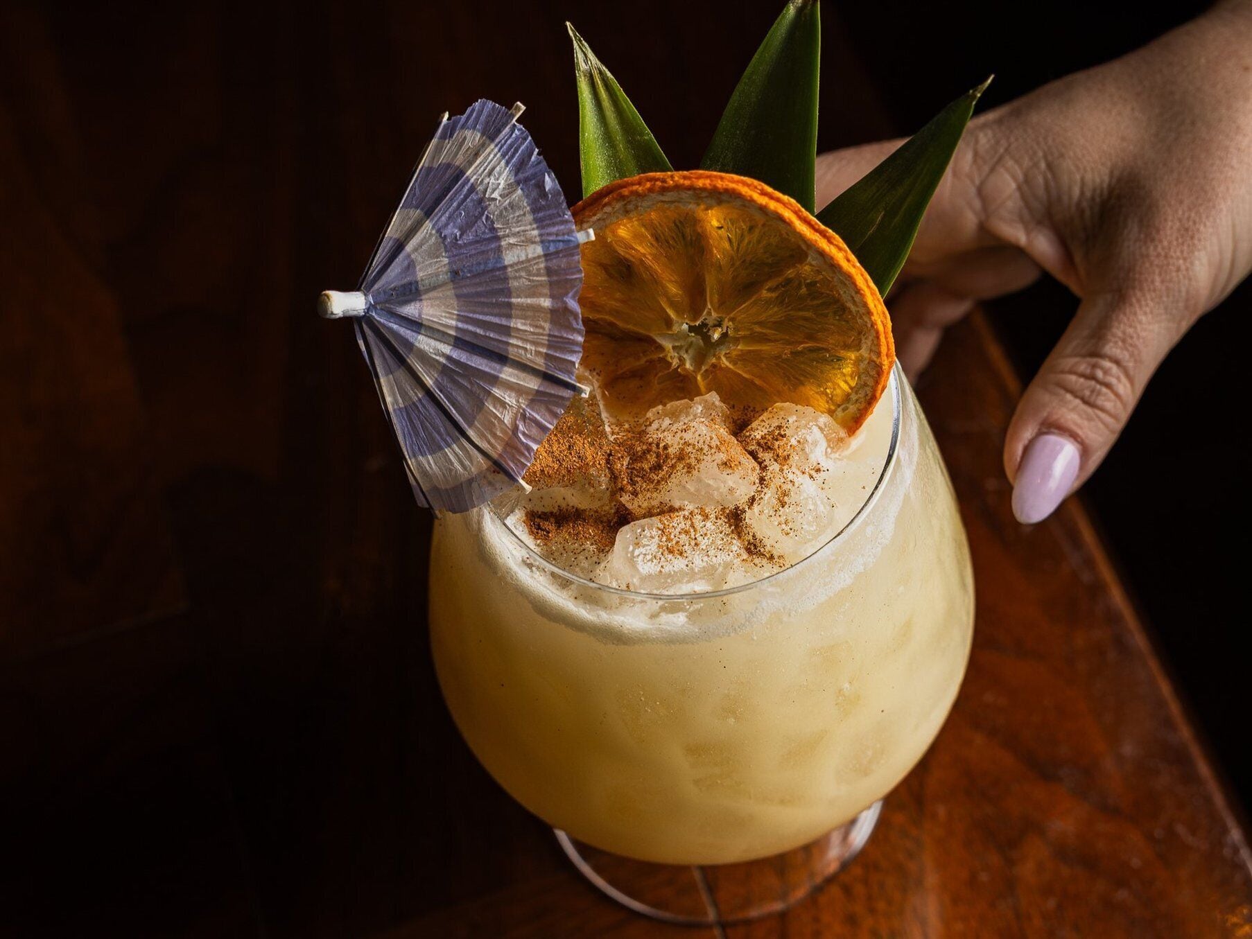 A cocktail with ice, a slice of orange, and a small umbrella on a glass.