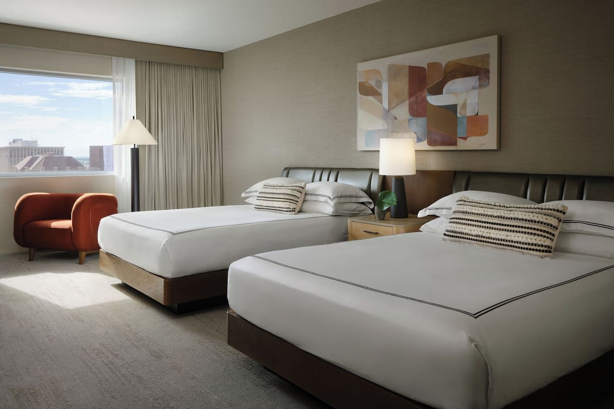 A hotel room with two beds, a chair, a lamp, and a city view.