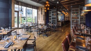 Un restaurante amplio con mesas y sillas de madera, ventanas grandes y lámparas colgantes.