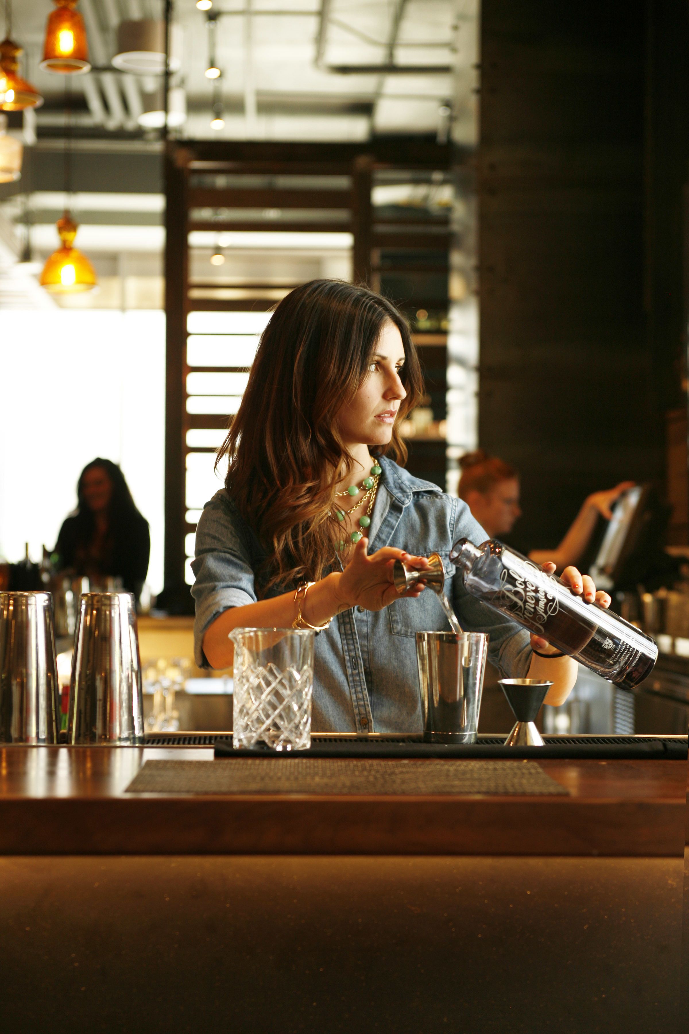 A bartender pouring a drink at a bar with a modern industrial design, including exposed ductwork and hanging pendant lights.