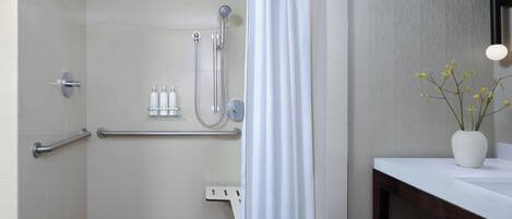 Un baño moderno con ducha, barras de apoyo, una cortina blanca y un lavamanos con un jarrón de flores.