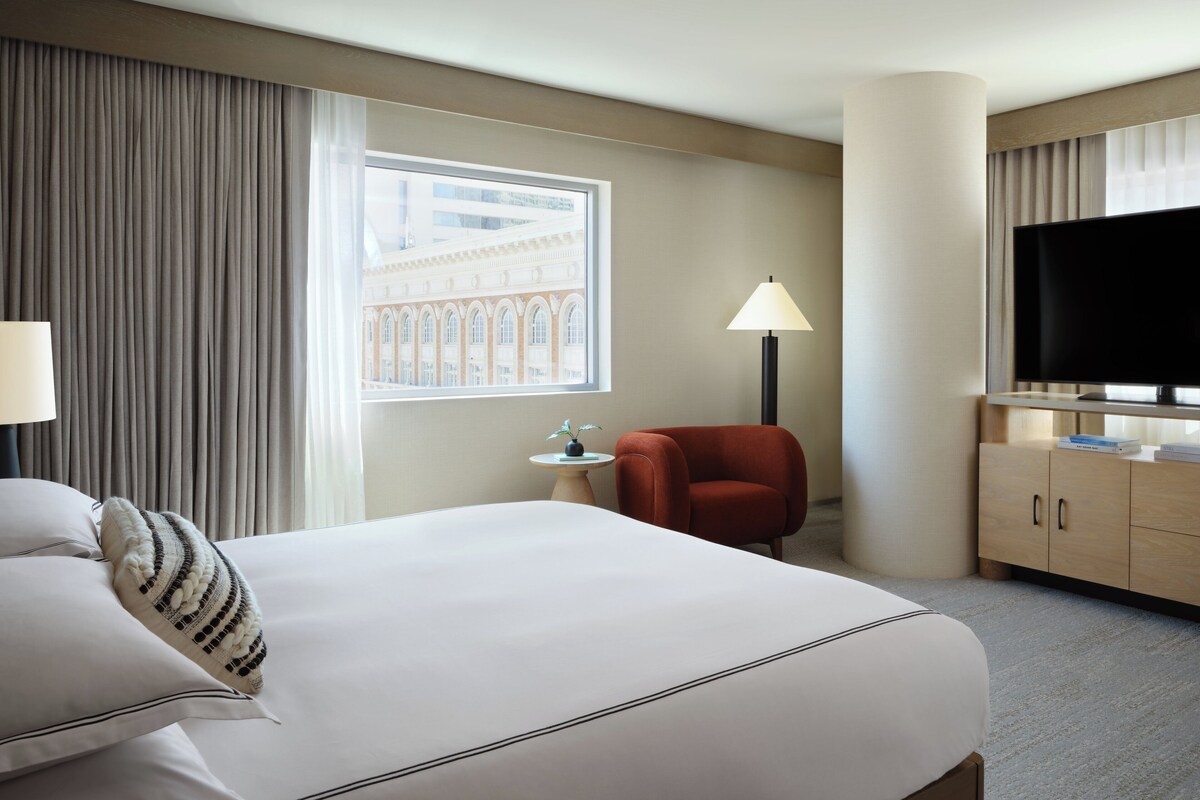A hotel room with a large bed, a red armchair, a small table with a lamp, a TV on a stand, and a view of a building through the window.