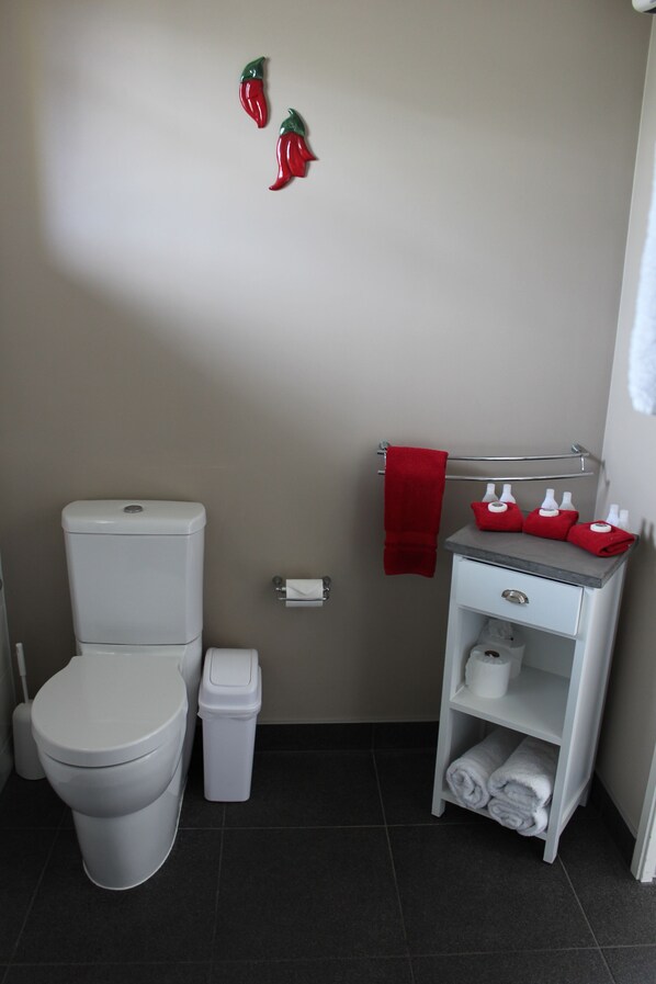 A bathroom with a toilet, a trash bin, a towel rack with a red towel, and a shelf with toiletries. - Chalet Queenstown (Queenstown)