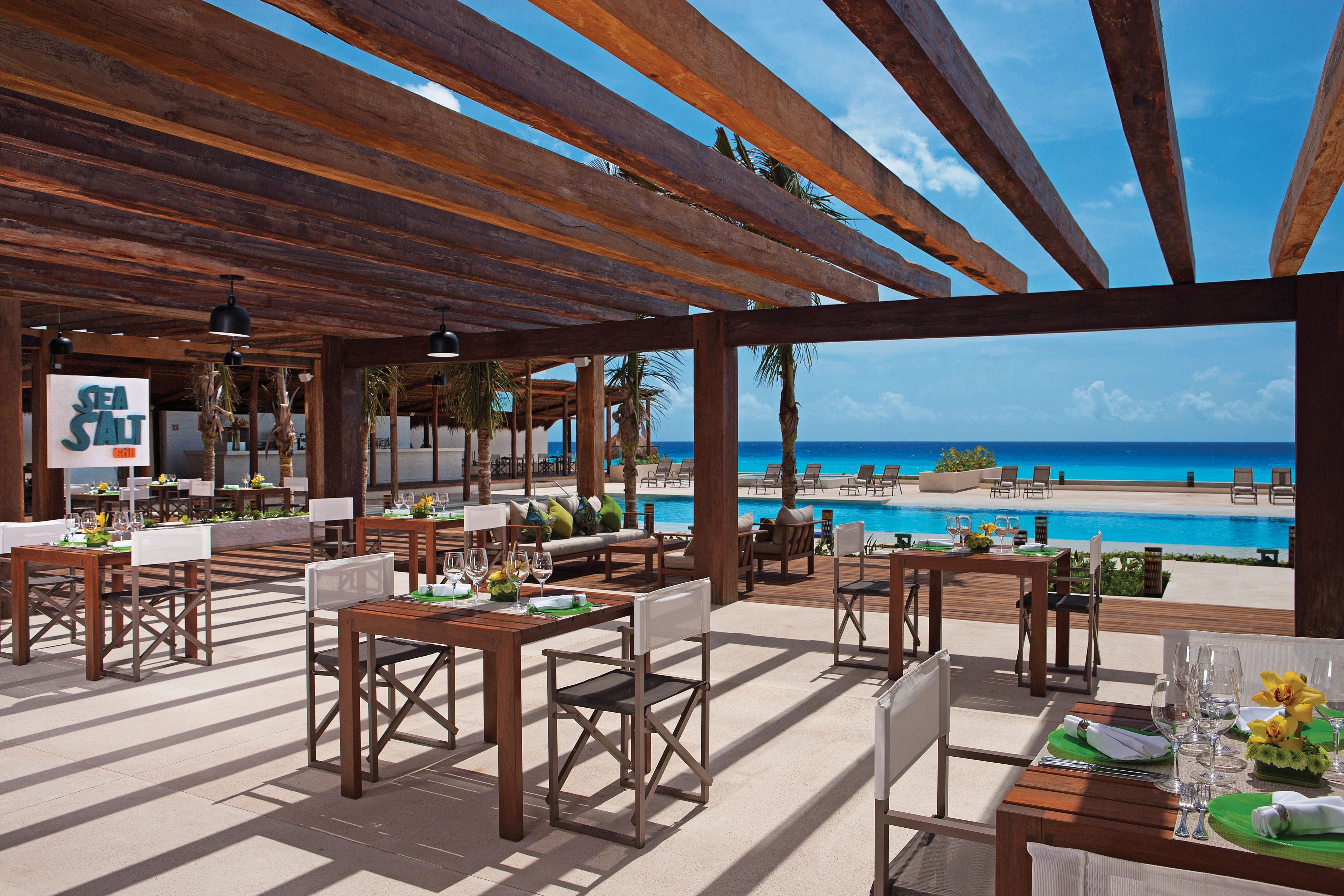 An outdoor dining area with wooden tables and chairs, overlooking a pool and beach.