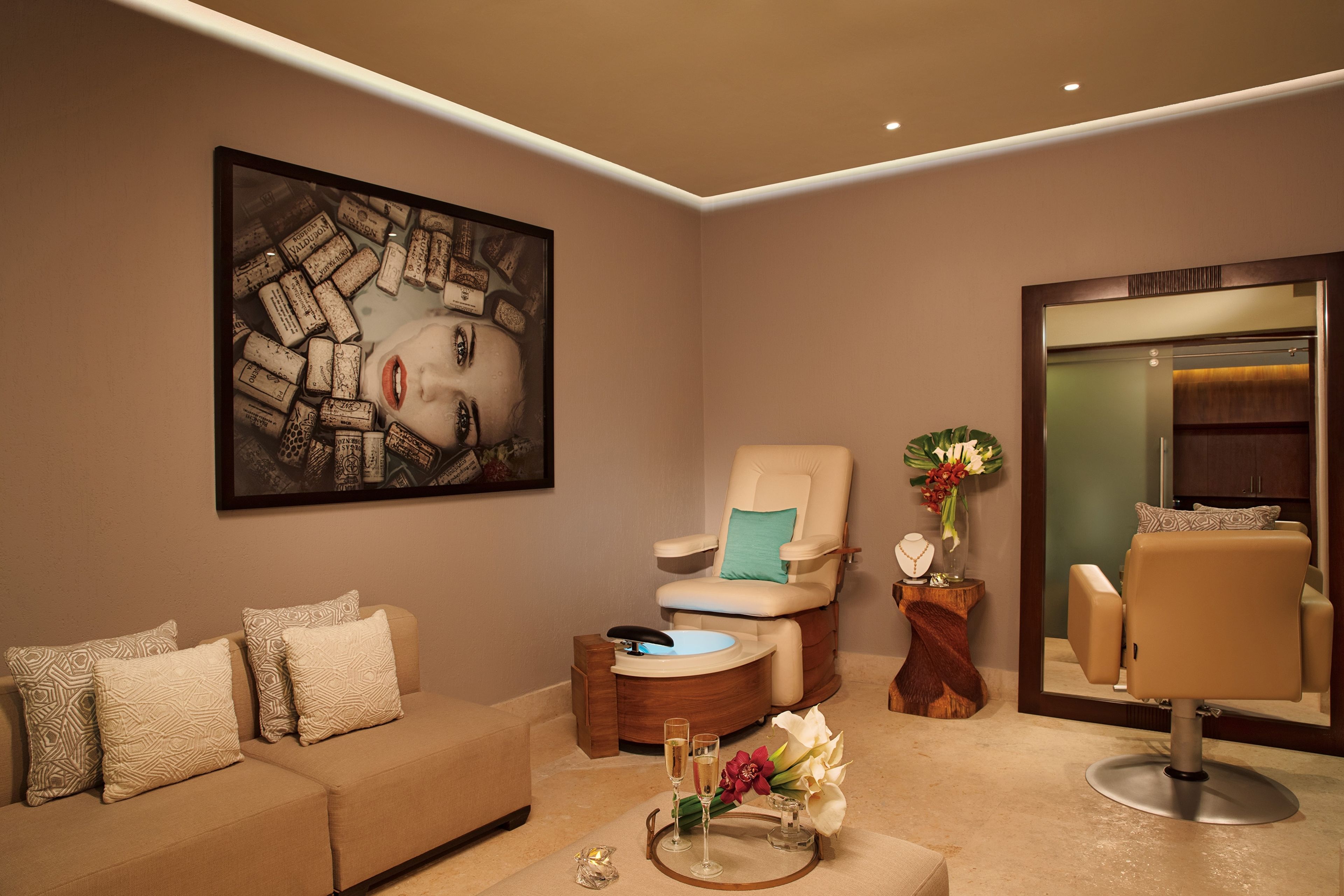 Een moderne kamer met een beige bank, een relax fauteuil, een groot kunstwerk, een spiegel en een bijzettafeltje met bloemen en glazen.