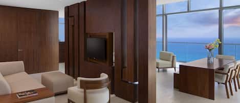 A modern living room with a flat-screen TV, a sofa, a chair, a dining table with chairs, and a view of the ocean through large windows.