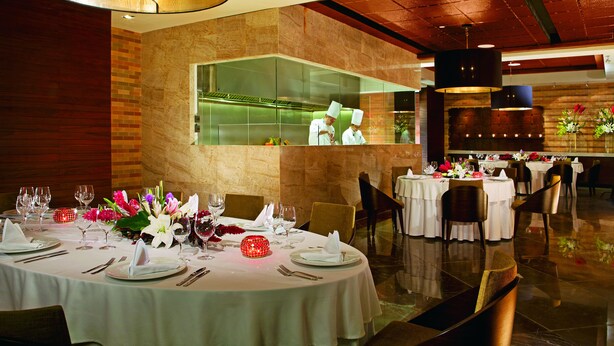 Un restaurante con mesas redondas dispuestas para comer, un interior moderno y vistas a la cocina.