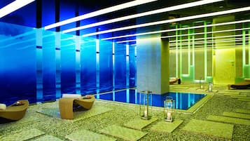 Un área de piscina interior moderna con agua azul cristalina, sillas de descanso e iluminación ambiental.