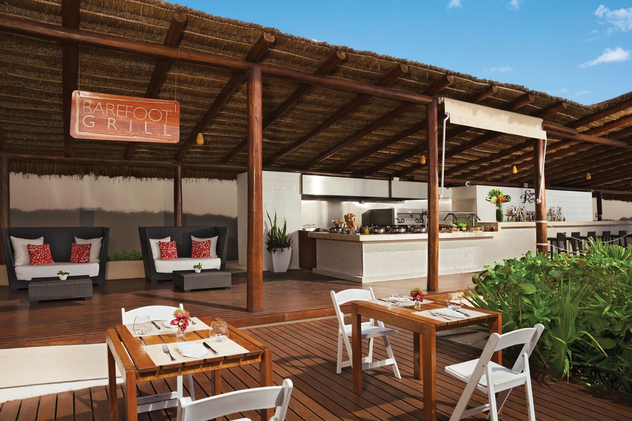An outdoor dining area with wooden tables and chairs, a bar, and a sign that reads 'BARBEFOOT GRILL'.