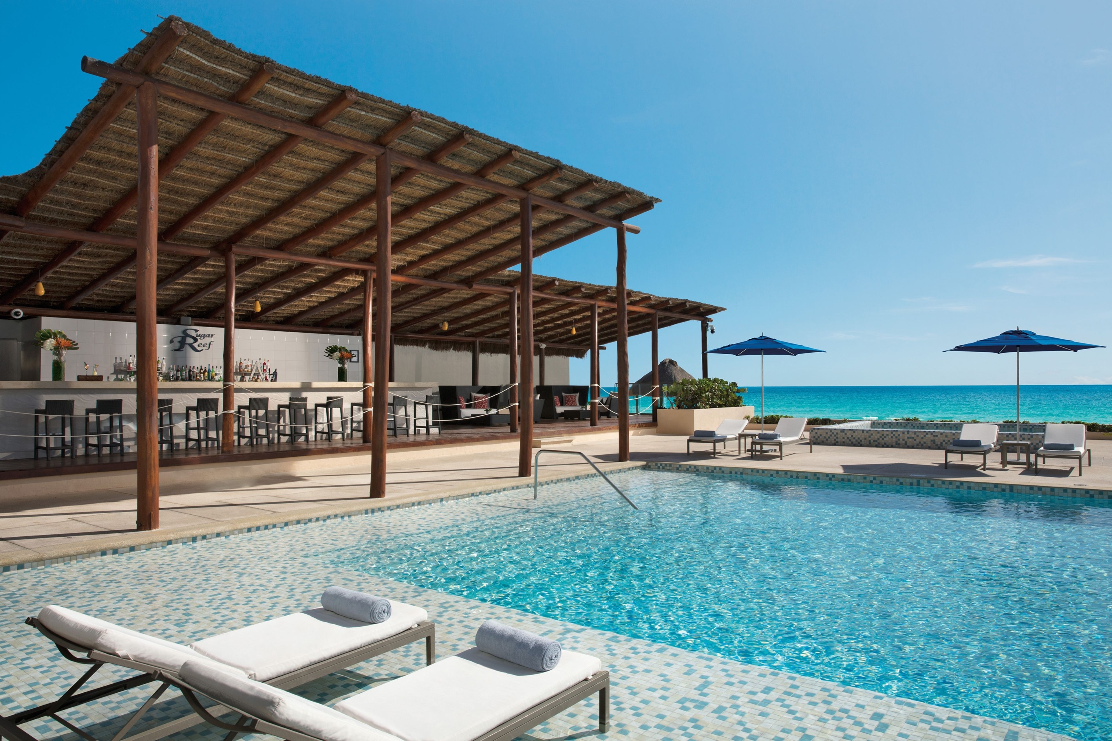 A pool area with lounge chairs and umbrellas, a bar, and a view of the ocean.