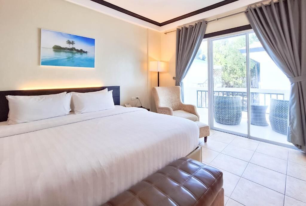 Quarto de hotel com uma cama grande, uma cadeira, varanda com móveis de vime e um quadro na parede.