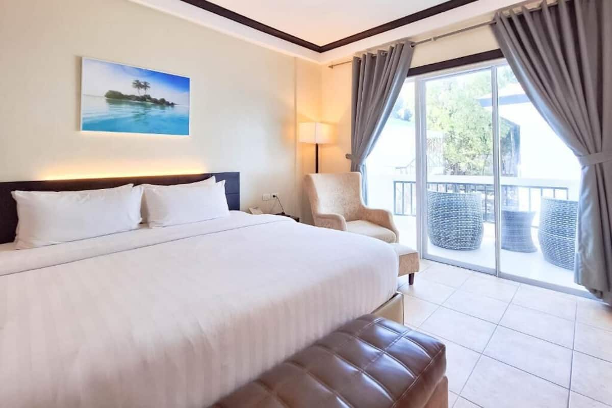 Ein Hotelzimmer mit einem großen Bett, einem Sessel, einem Balkon mit Korbmöbeln und einem Gemälde an der Wand.