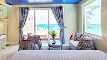 A hotel room with a bed, two wicker chairs, a small table with a plant, and a view of the ocean through a sliding door.