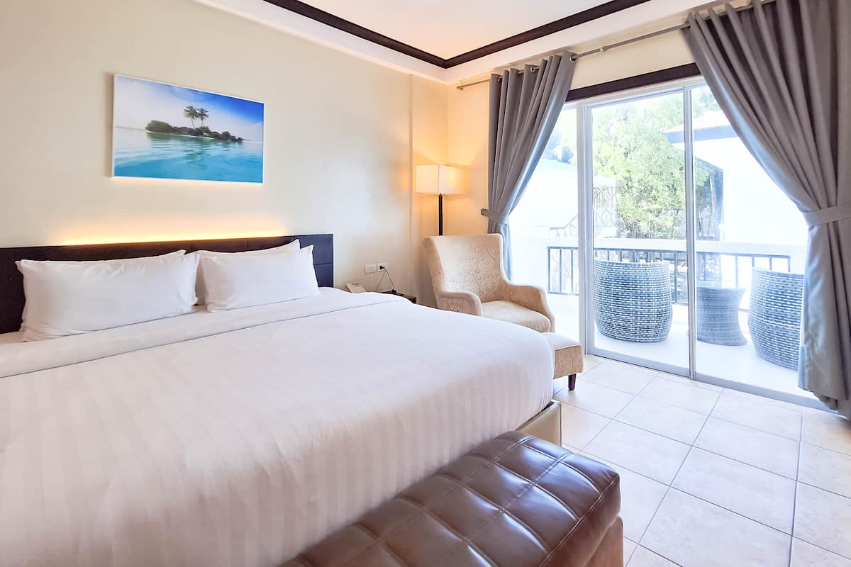 Ein Hotelzimmer mit einem großen Bett, einem Sessel, einem Balkon mit Korbmöbeln und einem Gemälde mit tropischer Szenerie.