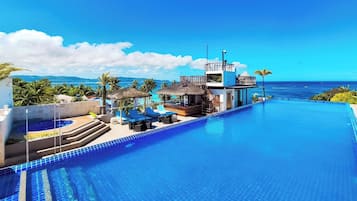 A resort pool with a view of the ocean, a lounge area with umbrellas, and a building with a balcony overlooking the water.