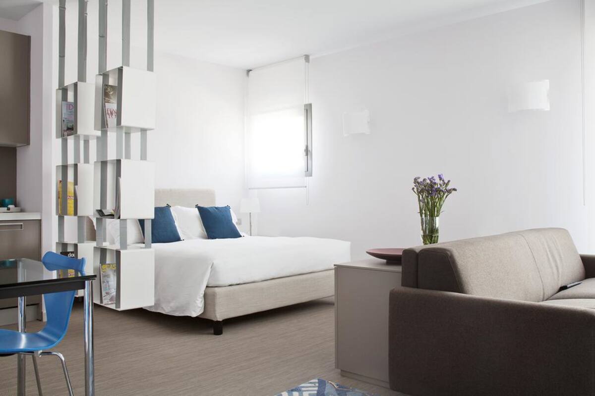 A modern hotel room with a bed, a sofa, a desk, and a chair. There is a bookshelf and a vase with flowers.