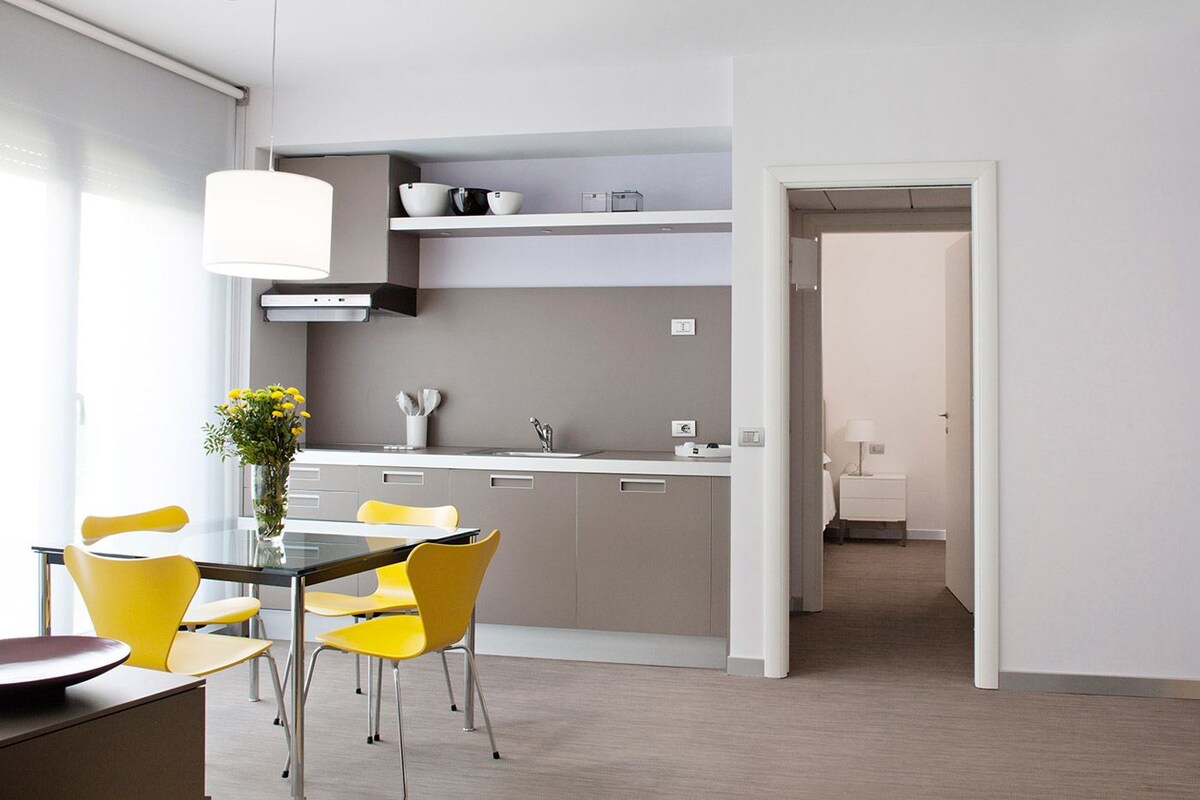 Una cocina moderna con zona de comedor, que presenta una mesa de vidrio y sillas amarillas, una lámpara colgante blanca y vistas al cuarto contiguo.