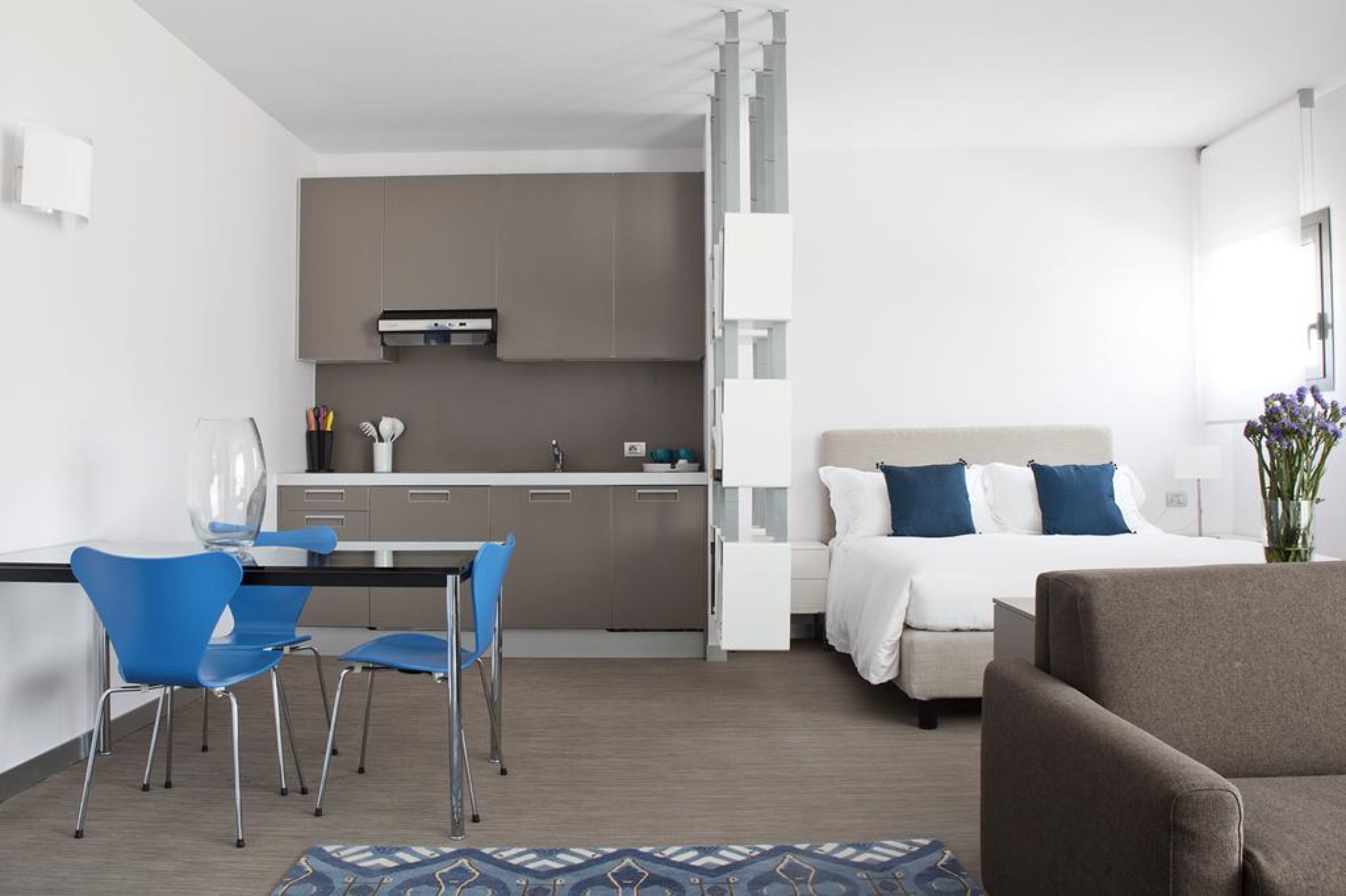 Una habitación de hotel moderna con kitchenette, un comedor con sillas azules, una cama con almohadas blancas y azules, y un sillón marrón.