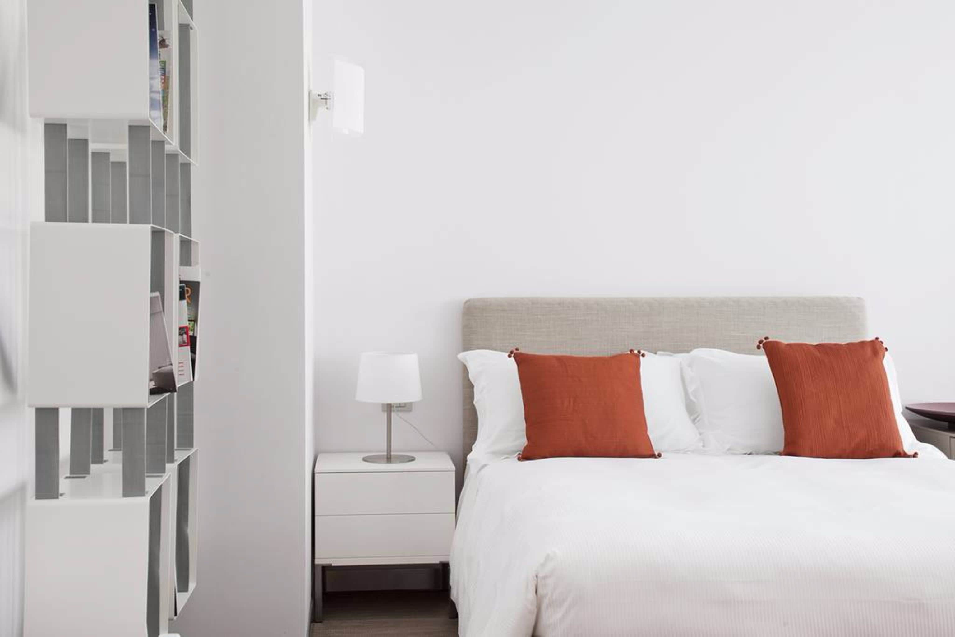 Ein Schlafzimmer mit einem Bett, weißen Kissen, einem Nachttisch, einer Lampe und einer weißen Regaleinheit.