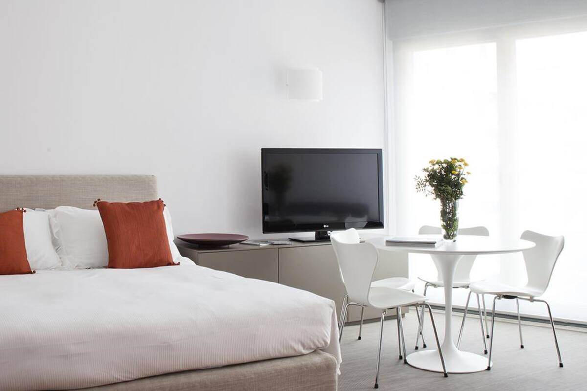 A modern hotel room with a bed, a TV, a dining table with chairs, and a window with natural light.