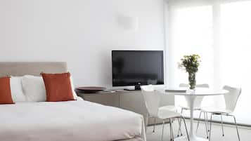 Quarto de hotel moderno com cama, televisão, mesa de jantar com cadeiras e janela com luz natural.