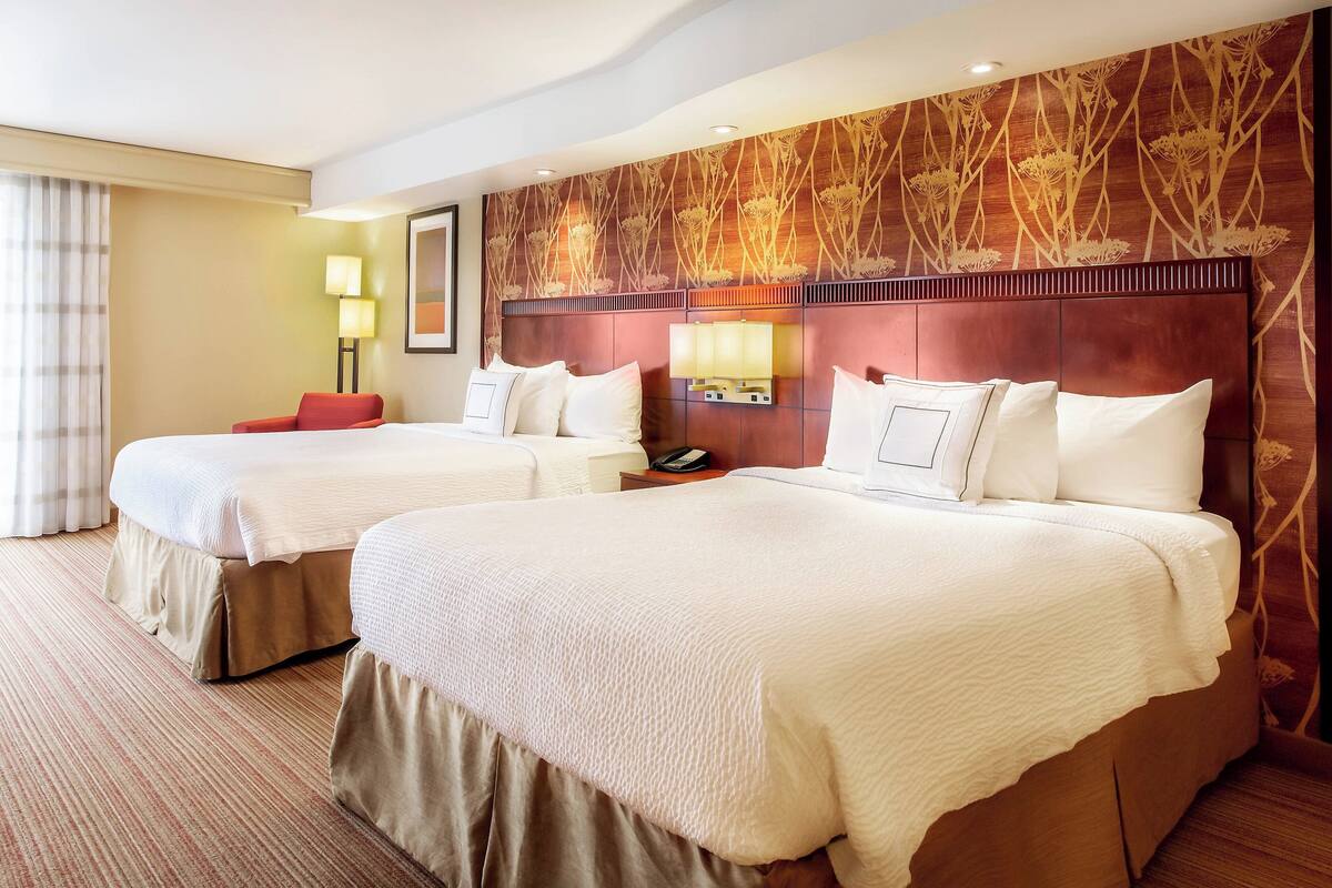 Habitación de hotel con dos camas, una silla roja, una mesita de noche con lámpara y un panel decorativo en la pared.