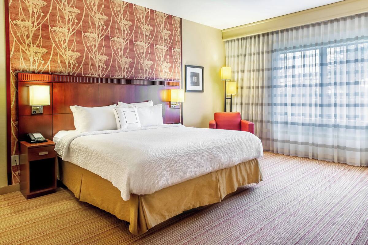 Habitación de hotel con una cama grande, una mesita de noche con teléfono, dos lámparas de noche, una silla roja y un ventanal con cortinas.