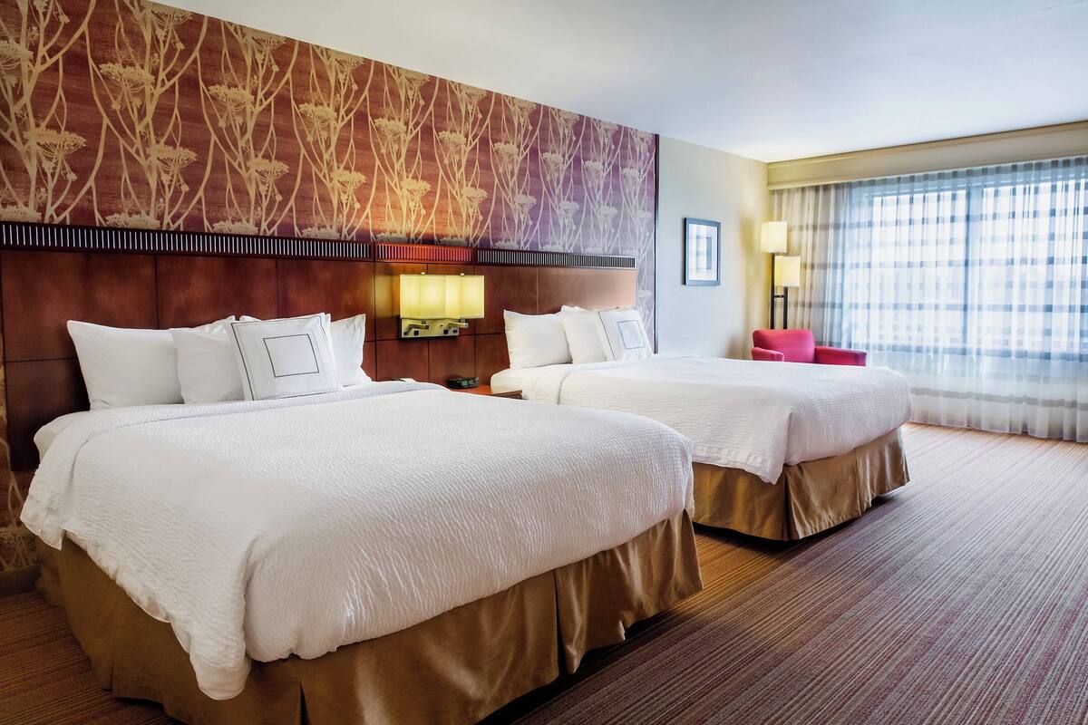 Habitación de hotel con dos camas, un cabecero grande estampado con motivos florales, una mesita de noche con lámpara y vistas al exterior a través de un ventanal amplio.