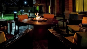 Un espace extérieur aménagé en terrasse, avec une table ronde entourée de chaises en osier, un foyer au centre et un mur de briques en arrière-plan.