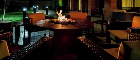An outdoor patio area with a round table surrounded by wicker chairs, a fire pit in the center, and a brick wall in the background.
