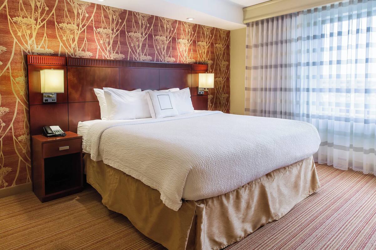 Habitación de hotel con una cama grande, dos mesitas de noche con lámparas, un teléfono sobre la mesa de la izquierda y una ventana con cortinas translúcidas.