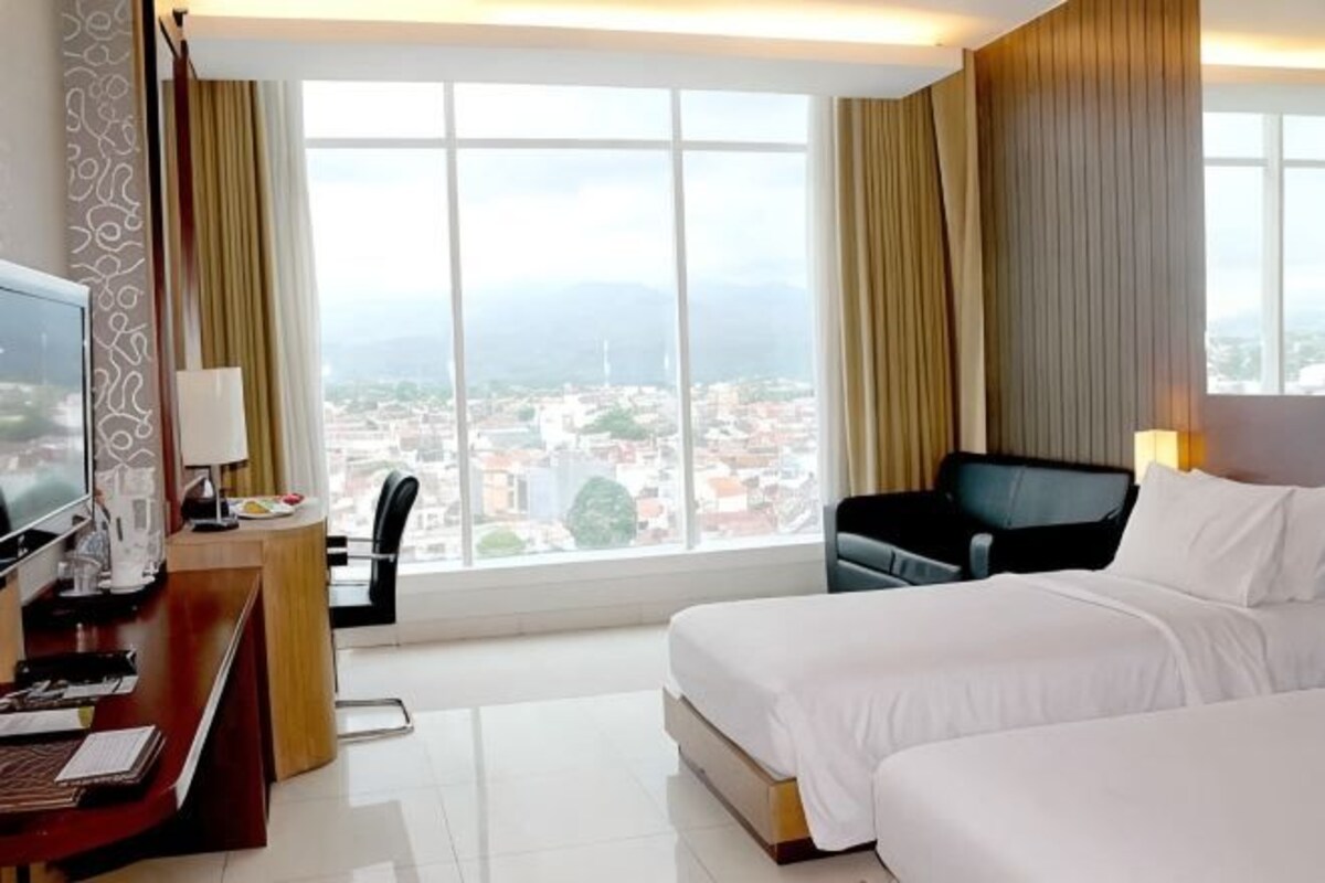 Habitación de hotel con cama, escritorio con televisor, una silla y vista a una ciudad a través de amplias ventanas.